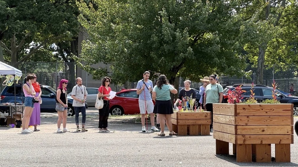 Diverse group of visitors touring a flower garden on a sunny day, standing in paved lot near raised beds of different heights. Some visitors have canes and walkers.